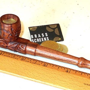 May include: A carved wooden pipe with a barrel-shaped bowl and a long stem. The bowl has a brass screen inside. A ruler shows measurements in inches. A small black card reads "BRASS SCREENS".