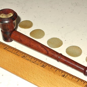 May include: A wooden smoking pipe with a brown and red finish. The pipe is about 7 inches long and has a bowl with a metal screen. The pipe is sitting on a wooden ruler with markings in inches. There are five round metal screens next to the pipe.