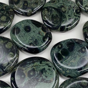 May include: A collection of polished, oval-shaped stones. The stones have a dark green base color with swirling patterns of lighter green and black. The stones are smooth and reflective, with a natural, organic appearance.