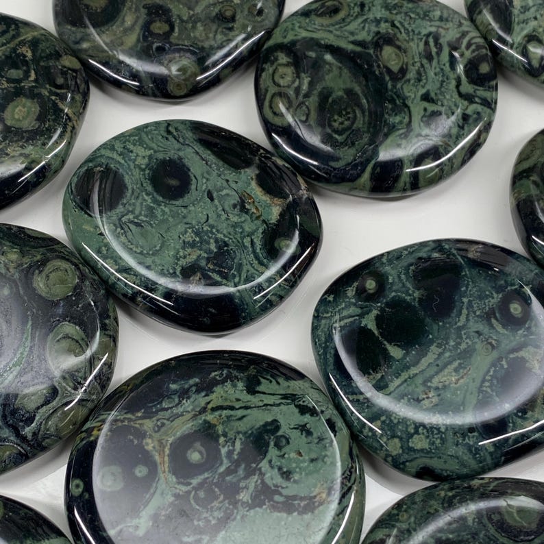 May include: A collection of polished, oval-shaped stones. The stones have a dark green base color with swirling patterns of lighter green and black. The stones are smooth and reflective, with a natural, organic appearance.