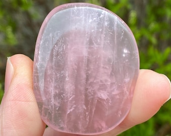 May include: A smooth, oval-shaped rose quartz stone. The stone is a soft pink color with translucent areas and subtle internal veining. The stone is held in a hand against a blurred green background.