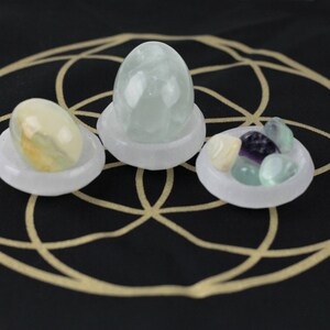 May include: Three small, white, shallow bowls each hold a different collection of stones. One bowl has a light green egg-shaped stone, another has a collection of small, colorful stones, and the third has a yellow and green stone. The bowls are on a black surface with a gold geometric design.