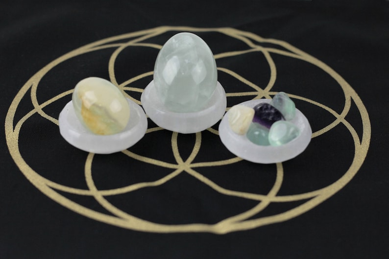 May include: Three small, white, shallow bowls each hold a different collection of stones. One bowl has a light green egg-shaped stone, another has a collection of small, colorful stones, and the third has a yellow and green stone. The bowls are on a black surface with a gold geometric design.