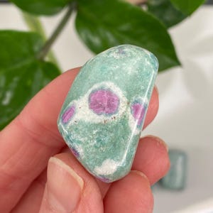 May include: A polished gemstone with a light teal base color and vibrant pink inclusions. The stone has a smooth, rounded shape and is held in a hand, with a green plant in the background. The pink inclusions are scattered throughout the stone.