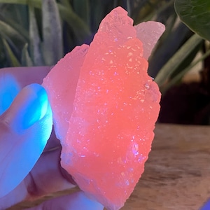 Peut inclure: Gros plan sur un amas de cristaux roses à surface rugueuse et texturée. Le cristal est éclairé d'une douce lueur rose, avec des reflets bleus. Le cristal est tenu dans une main.