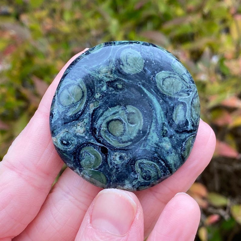 May include: A round, polished stone with a dark green and teal pattern. The stone has a swirling, eye-like design, with lighter green circles against a darker background. The stone is held in a hand, with a blurred natural background.