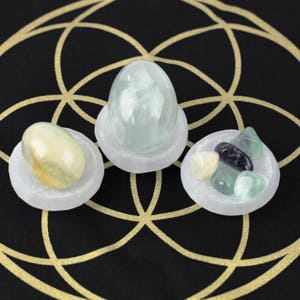 May include: Three selenite bowls each holding different crystals. One bowl contains a large, light green crystal. Another holds a mix of small, colorful stones. The third bowl has a yellow and white stone. The background is black with a gold geometric design.