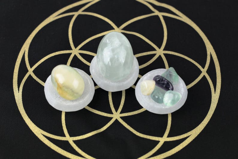 May include: Three selenite bowls each holding different crystals. One bowl contains a large, light green crystal. Another holds a mix of small, colorful stones. The third bowl has a yellow and white stone. The background is black with a gold geometric design.