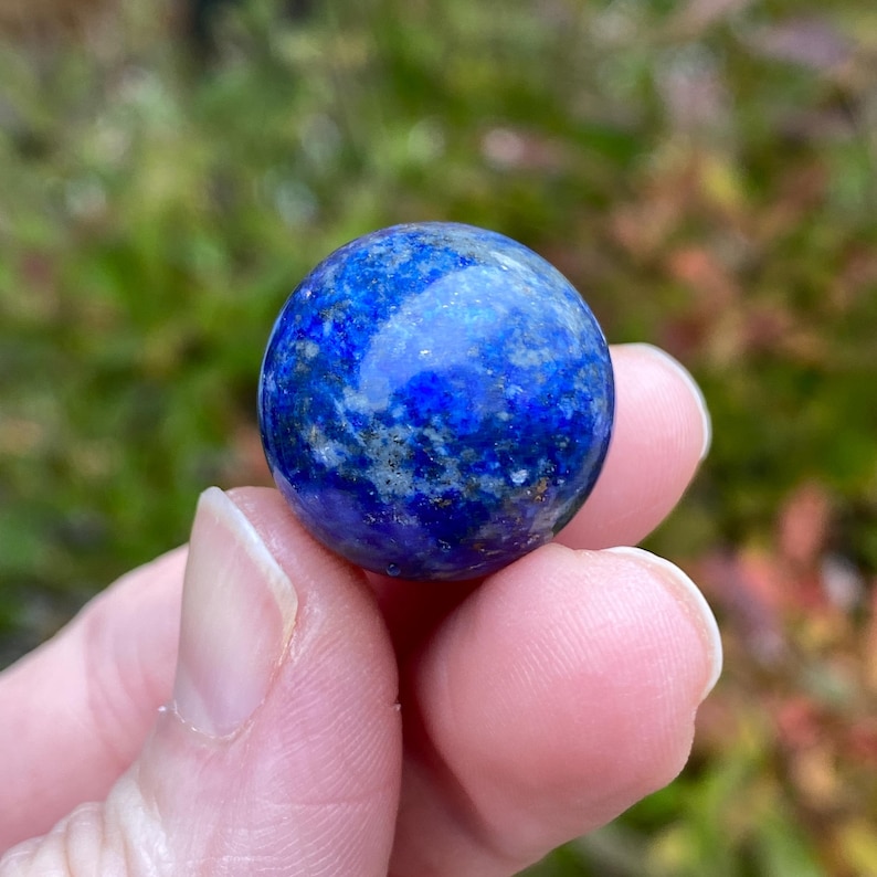 May include: A polished, spherical lapis lazuli gemstone. The deep blue stone has flecks of gold and white throughout its surface. The gemstone is held in a person's hand against a blurred green background.
