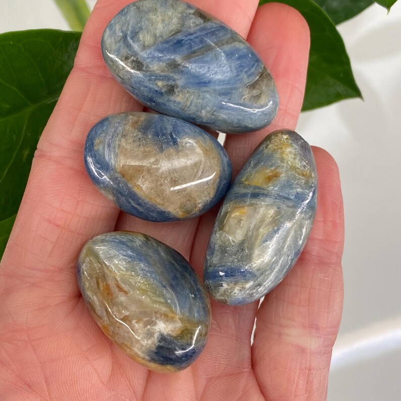 Puede incluir: Cuatro piedras de cianita azul pulidas con inclusiones de color beige y blanco se sostienen en una mano. Las piedras ovaladas tienen un acabado liso y brillante. La cianita tiene un aspecto natural y terroso.