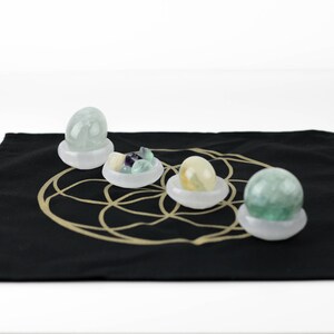 May include: Four polished gemstone spheres and clusters in white bowls, displayed on a black cloth with a gold geometric design. The gemstones are various colors, including green, yellow, and purple.