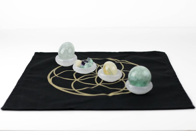 May include: Four polished gemstone spheres and clusters in white bowls, displayed on a black cloth with a gold geometric design. The gemstones are various colors, including green, yellow, and purple.