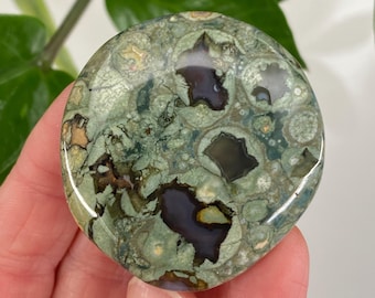 May include: A round, polished gemstone with a mottled pattern of green, brown, and dark purple. The stone has a smooth, glossy surface and is held in a hand. The gemstone's unique coloration and texture are highlighted.