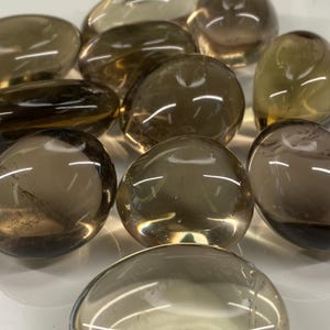 May include: A collection of smooth, polished smoky quartz stones. The stones vary in shades of brown and tan, with a translucent quality. The rounded shapes and glossy surfaces reflect light, creating highlights. These stones are often used for meditation and healing.