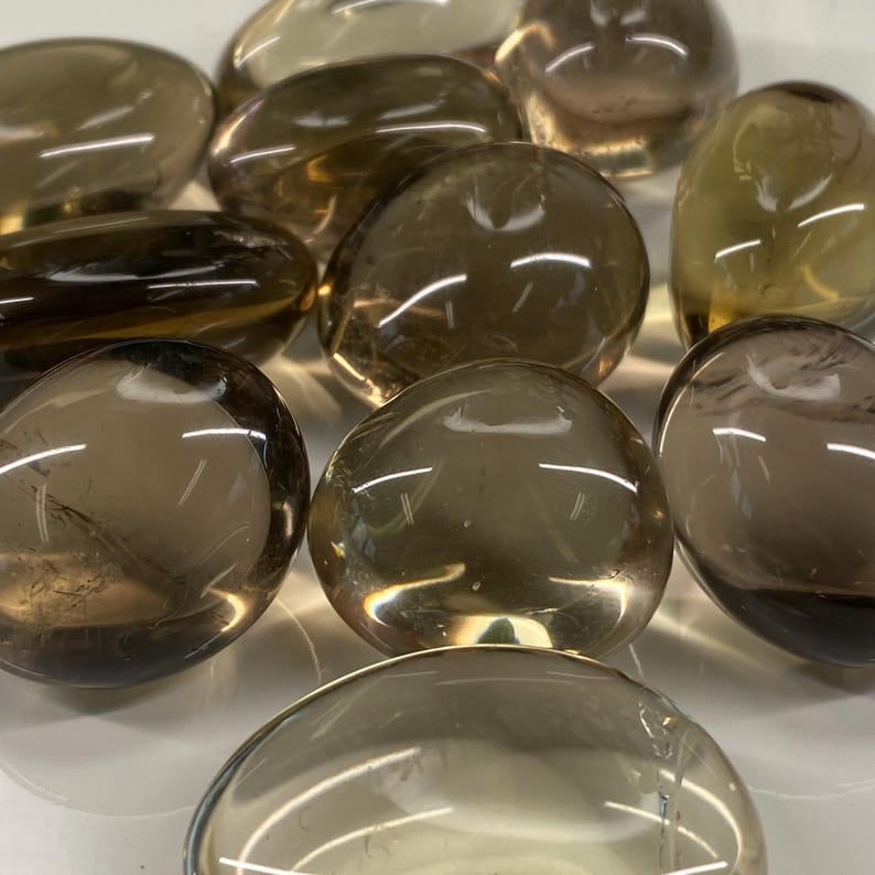 May include: A collection of smooth, polished smoky quartz stones. The stones vary in shades of brown and tan, with a translucent quality. The rounded shapes and glossy surfaces reflect light, creating highlights. These stones are often used for meditation and healing.