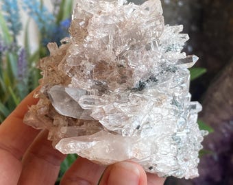 May include: A cluster of clear quartz crystals with a light brown hue. The crystals are clustered together, with some individual points visible. The quartz has a natural, raw appearance, with some dark green and black inclusions.