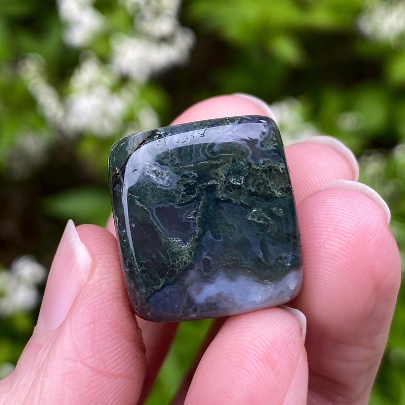 Puede incluir: Una piedra de &aacute;gata musgosa pulida, de forma cuadrada. La piedra presenta una superficie transl&uacute;cida y brillante con patrones verdes, blancos y azules. La piedra se sostiene en una mano, con un fondo verde borroso.