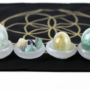 May include: Four selenite bowls holding various crystals and stones. The bowls are white and sit on a black surface with a gold geometric design. The crystals include green, yellow, and clear egg-shaped stones and a green sphere.