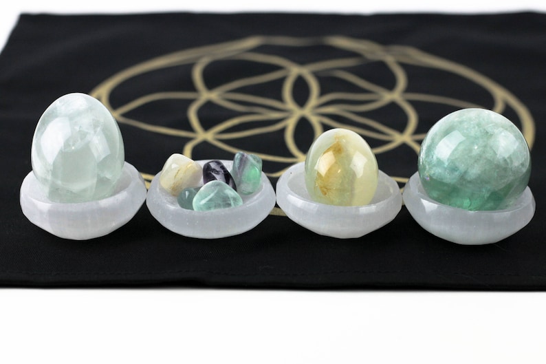 May include: Four selenite bowls holding various crystals and stones. The bowls are white and sit on a black surface with a gold geometric design. The crystals include green, yellow, and clear egg-shaped stones and a green sphere.