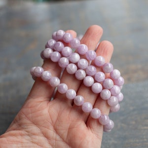 May include: A strand of pale lavender gemstone beads, likely amethyst, with a smooth, polished finish. The beads are round and uniform in size, creating a consistent look. The strand is coiled in a hand, showcasing the colour and texture of the beads.