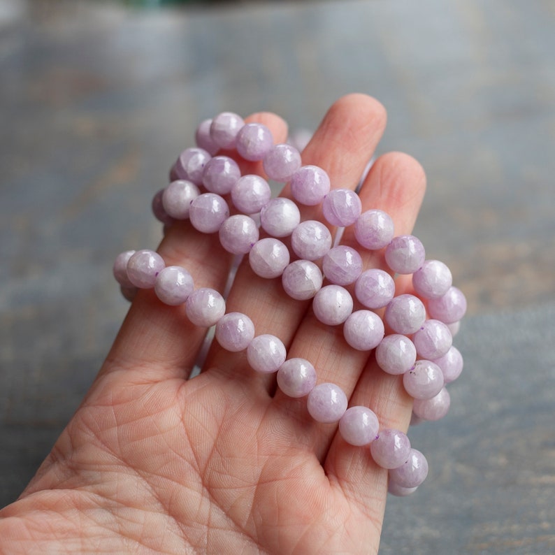 May include: A strand of pale lavender gemstone beads, likely amethyst, with a smooth, polished finish. The beads are round and uniform in size, creating a consistent look. The strand is coiled in a hand, showcasing the colour and texture of the beads.