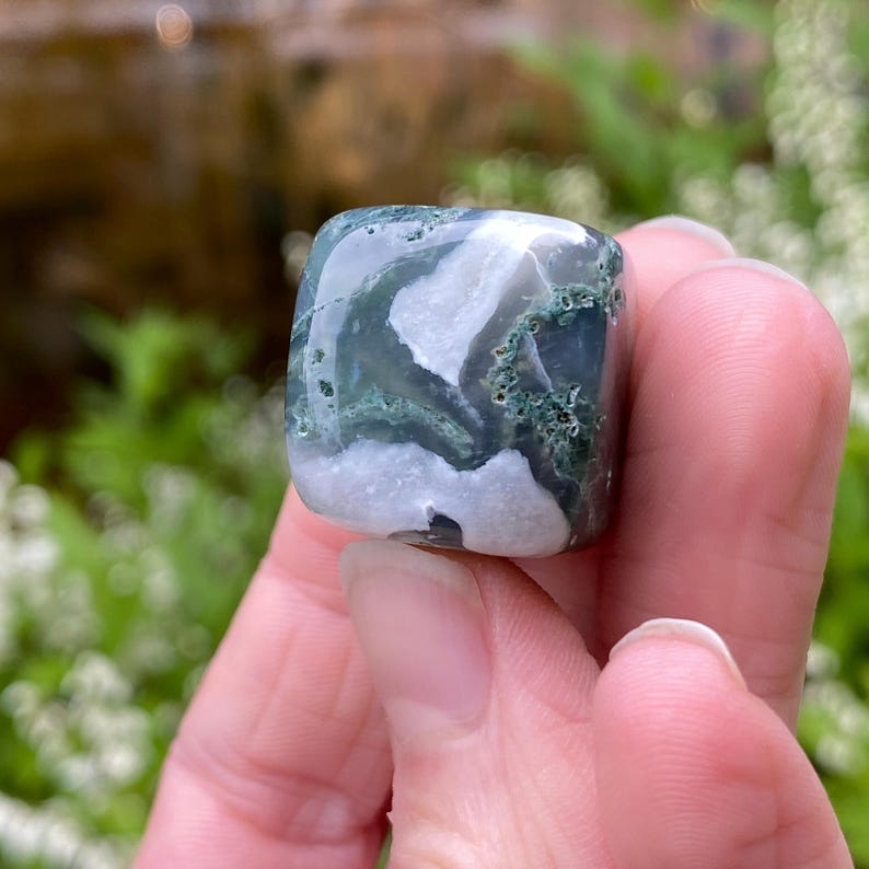 Puede incluir: Una piedra preciosa de &aacute;gata musgosa de forma cuadrada con una superficie pulida. La piedra presenta una mezcla de patrones blancos, verdes y grises. La piedra preciosa se sostiene en una mano, con un fondo verde borroso.
