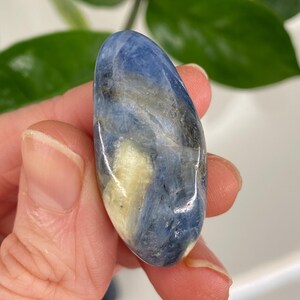 Puede incluir: Una gema pulida, de forma ovalada, con una mezcla de colores azul, blanco y crema. La piedra tiene una superficie lisa y brillante, sostenida en una mano. Los patrones naturales crean un efecto veteado &uacute;nico. Mide aproximadamente 5 cm.