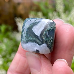 Puede incluir: Una piedra preciosa de &aacute;gata musgosa pulida, de forma cuadrada. La piedra presenta una mezcla de patrones blancos transl&uacute;cidos, verdes oscuros y grises. La &aacute;gata se sostiene en una mano, con un fondo verde borroso.