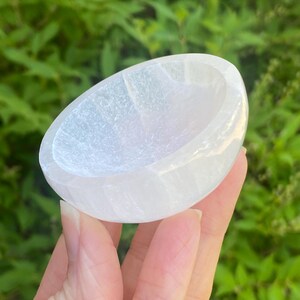 May include: A white selenite crystal bowl, oval-shaped with a smooth, polished exterior. The bowl has a shallow interior and is held in a hand against a backdrop of green foliage. The selenite has a translucent, almost pearlescent quality.