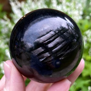 Peut inclure: Une boule de cristal noire polie et sphérique. La surface reflète la lumière, créant un aspect brillant. Le cristal est tenu dans une main, avec un arrière-plan vert et blanc flou.