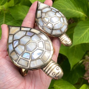 May include: Two turtle-shaped decorative objects crafted from polished brass and inlaid with iridescent, white, and cream-colored shell segments. Each turtle features a segmented shell pattern and a smooth, metallic head with etched eyes.