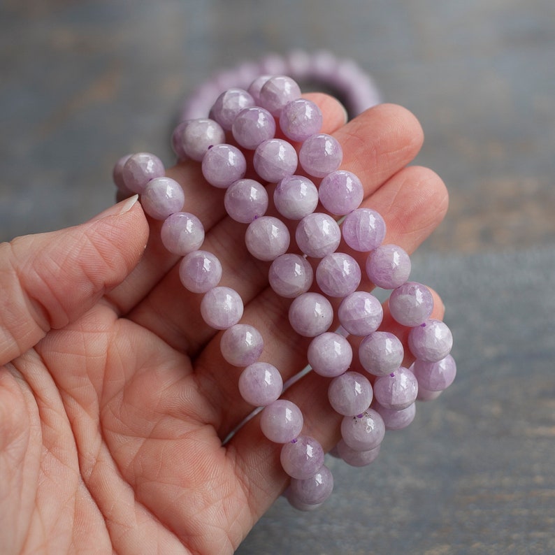 May include: A strand of lavender amethyst beads, with a smooth, polished finish. The beads are round and uniform in size, and they are strung on a clear elastic cord.