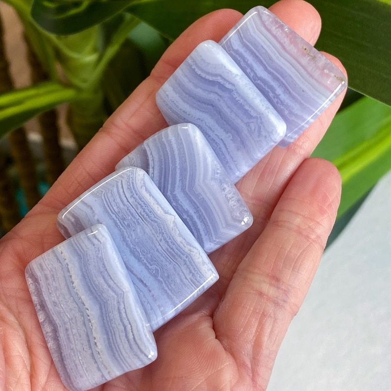 May include: Six polished blue lace agate stones, rectangular in shape, are displayed in a hand. The stones have a soft, banded appearance with shades of light blue and white. The stones are smooth and have a glossy finish.