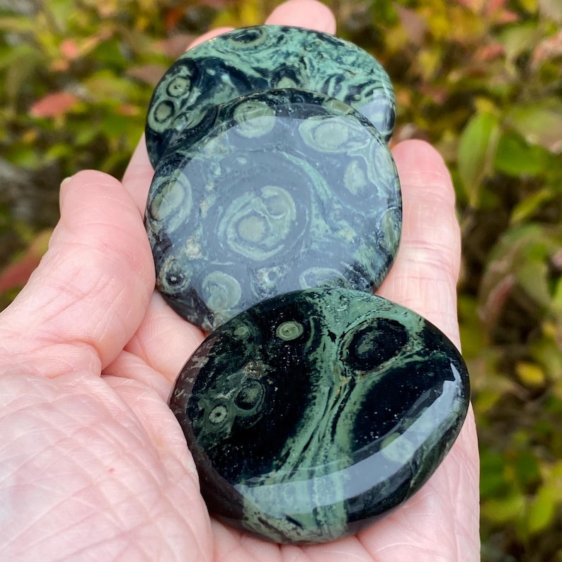 May include: Three polished, round stones with a dark green and black swirling pattern are held in a hand. The stones have a glossy finish and a unique, natural design. The background is blurred with green foliage.
