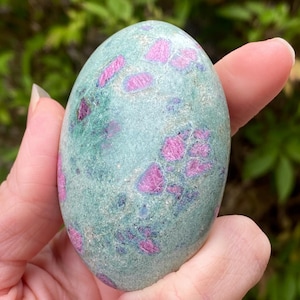 Peut inclure: Une pierre précieuse ovale polie, avec une base turquoise et des inclusions de rubis roses vives. La pierre est tenue dans une main, mettant en valeur sa texture lisse et sa beauté naturelle. Les cristaux de rubis sont regroupés.