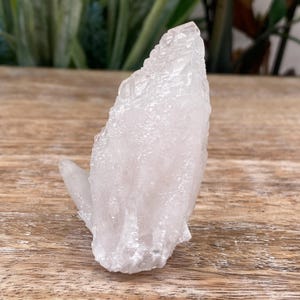Peut inclure: Un amas de cristaux de quartz clair avec une surface rugueuse et texturée. Le cristal est d'un blanc pâle et translucide et repose sur une surface en bois. Le cristal a un corps principal avec un cristal plus petit qui s'étend sur le côté.