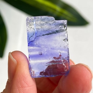 Puede incluir: Cristal rectangular translúcido con tonos morados y azules. El cristal, sostenido entre los dedos, muestra una textura rugosa y natural. La gema tiene una apariencia en capas, con estructuras internas visibles. La estética general es orgánica y terrosa.