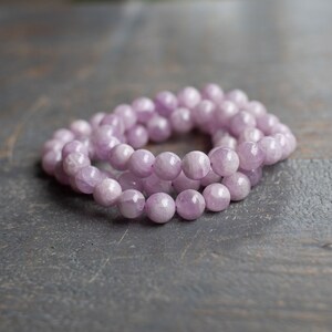 May include: A strand of lavender-coloured gemstone beads, likely amethyst, arranged in a circular shape on a grey textured surface.