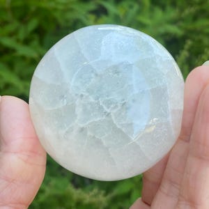 May include: A translucent, spherical crystal ball with a pale blue-green hue. The surface has a textured, fractured appearance, with subtle variations in color and light refraction. The crystal is held in a person's hand, with a blurred green background.