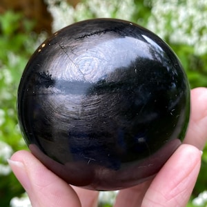 Peut inclure: Une sphère noire polie avec une surface réfléchissante. La sphère présente un motif circulaire et une fissure sur le dessus. La sphère est tenue dans la main d'une personne, avec un arrière-plan vert flou.