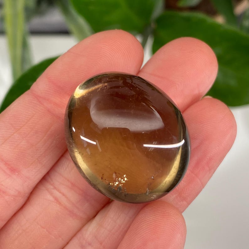 May include: A smooth, oval-shaped, translucent gemstone in shades of brown and amber. The polished stone has a glossy surface and is held in a hand, with a blurred green background.