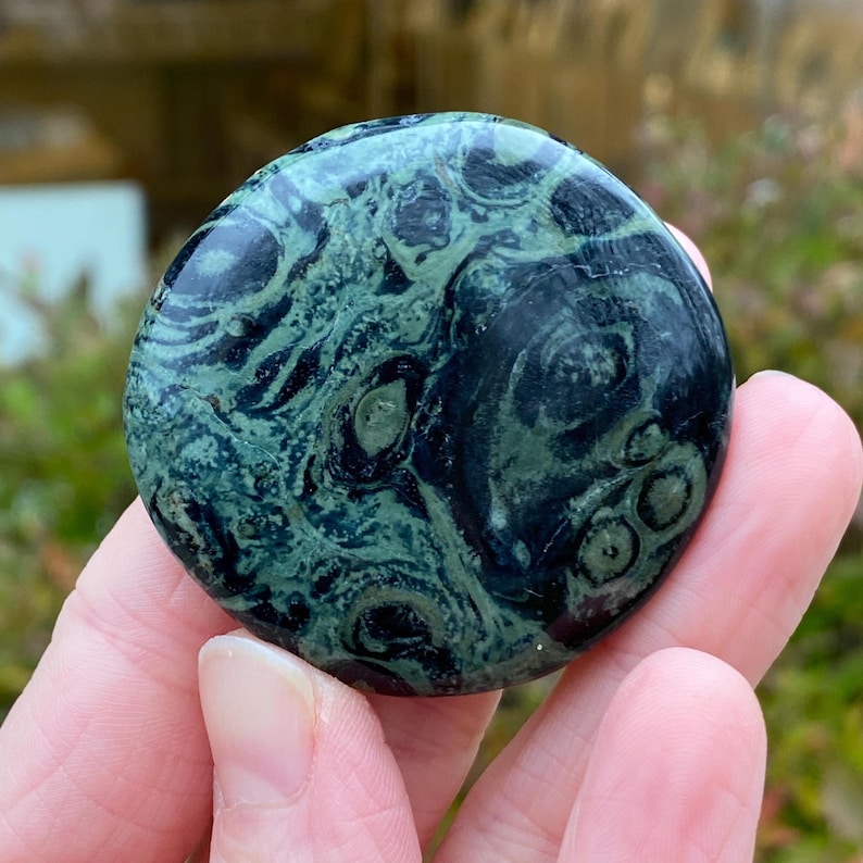 May include: A round, polished stone with a swirling pattern of dark green and black. The stone is held in a hand, showcasing its smooth, glossy surface and unique natural design. The stone's colors and patterns create a striking visual effect.