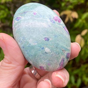 May include: A smooth, oval-shaped stone with a teal base color and scattered pink and blue inclusions. The stone is held in a hand, showcasing its polished surface and natural variations. The stone's size is approximately 3 inches in length.