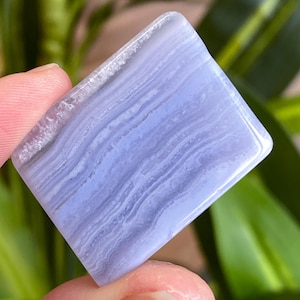 May include: A square, polished blue lace agate gemstone. The stone has a light blue hue with white and darker blue banding patterns. The edges are smooth and rounded. The gemstone is held between two fingers.