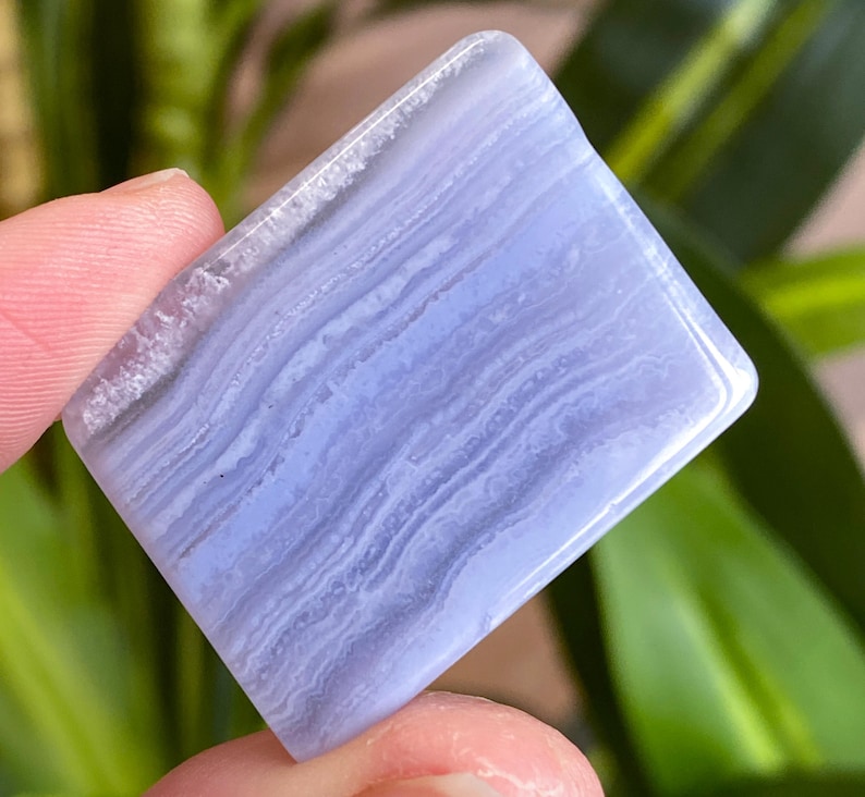 May include: A square, polished blue lace agate gemstone. The stone has a light blue hue with white and darker blue banding patterns. The edges are smooth and rounded. The gemstone is held between two fingers.