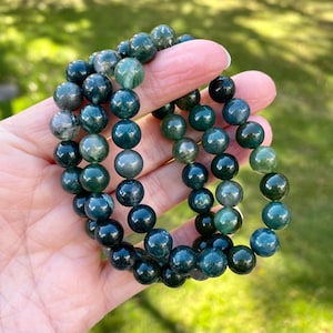May include: Three green moss agate beaded bracelets stacked on top of each other. The beads are round and have a dark green color with lighter green and white inclusions.