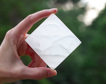 May include: A square, white decorative object with a textured surface is held in a hand. The object has a diamond shape and appears to be made of a translucent material. The background is a blurred green.