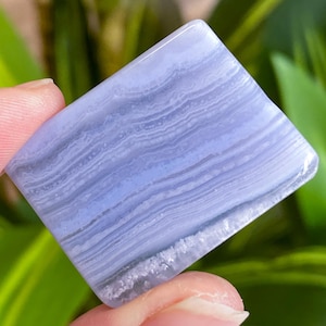 May include: A polished, rectangular blue lace agate gemstone. The stone displays a layered pattern of light and dark blue bands, with a smooth, glossy surface. The edges are slightly rounded, and the stone is held by a person's fingers.