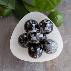May include: A white bowl filled with six black and white snowflake obsidian spheres. The spheres are smooth and polished, with a speckled pattern of white on black.