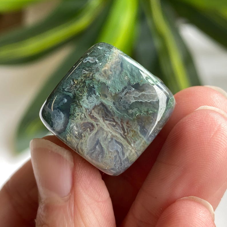 Puede incluir: Una gema pulida de forma cuadrada con una mezcla de tonos verdes, blancos y azules. La piedra tiene una cualidad transl&uacute;cida, revelando intrincados patrones internos. La gema se sostiene en una mano, con un fondo verde borroso.